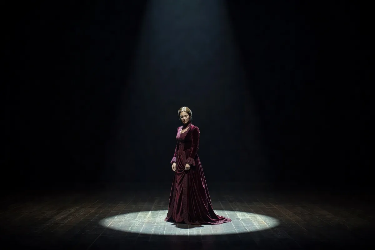 Comédienne en robe d'époque seule sur une scène sombre sous un projecteur