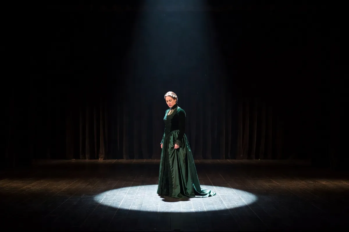 Comédienne en robe d'époque seule sur une scène sombre sous un projecteur
